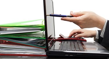 Ein Foto eines Buchhalters mit einem Laptop auf einem Tisch, links davon ein Stapel bunter Ordner. Der Buchhalter zeigt mit einem Stift auf den Bildschirm.