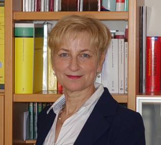Foto von Hannelore Bäuml, stehend vor einem Bücherregal, mit einem weißen Hemd und einem dunklen Blazer bekleidet.